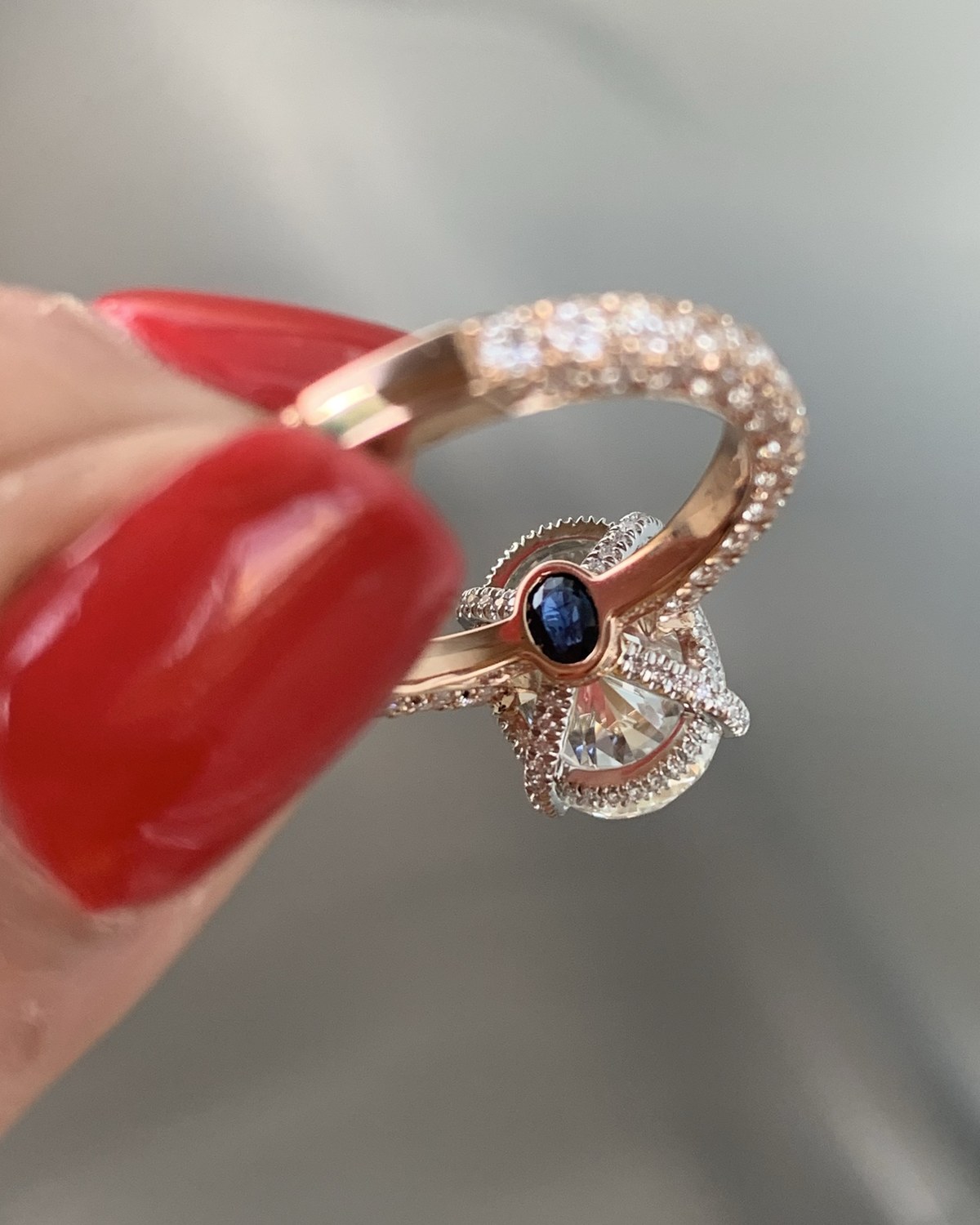 Warm rose gold bypass ring carrying a small oval blue stone on one side and a pavé diamond form on the other, held in the fingers