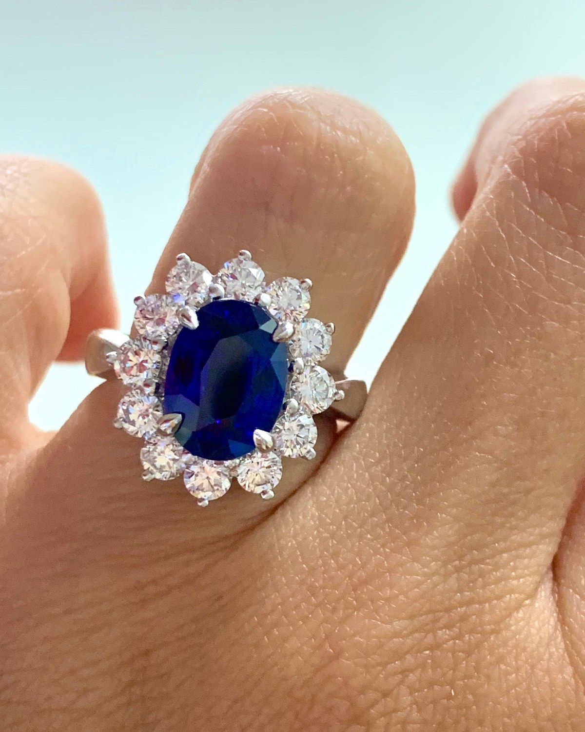 Deep blue oval center stone in a claw-set surround of round diamonds, worn on the hand in daylight