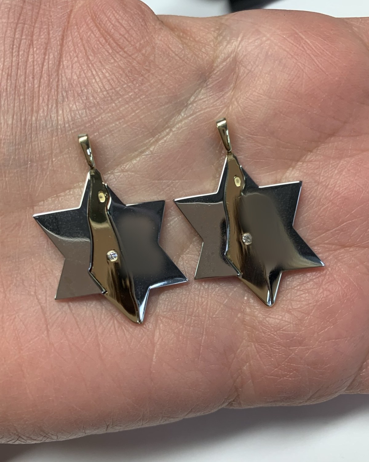 Two Star of David pendants in white and yellow gold, each with a raised ribbon panel whose engraved lettering is deliberately obscured, held in the palm