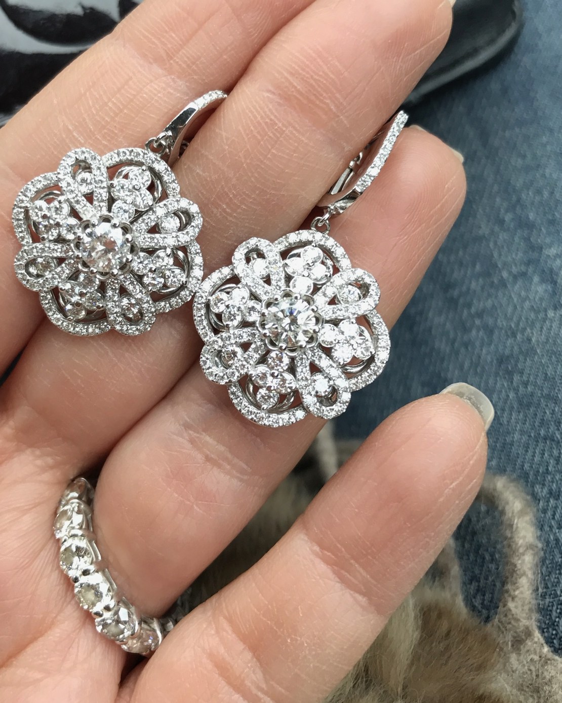 Matched pair of white gold floral diamond cluster drop earrings held in the fingers beside a diamond band